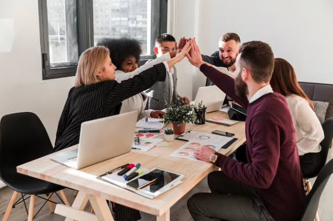 Equipe reunida ao redor de uma mesa de trabalho celebrando em conjunto, com os integrantes levantando as mãos em gesto de cooperação.