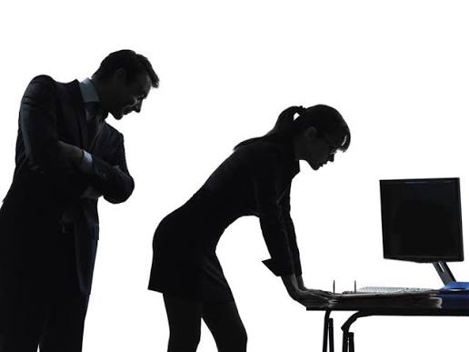 Silhueta de um homem observando uma mulher que trabalha diante de um computador em um escritório.