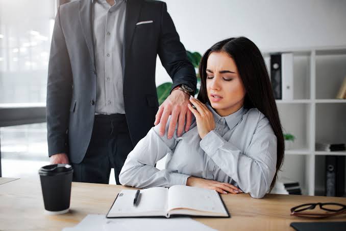 Mulher afasta a mão de um homem que toca seu ombro no ambiente de trabalho.