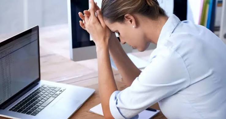 Mulher diante de um notebook, com as mãos apoiadas na testa e expressão de cansaço durante o trabalho.