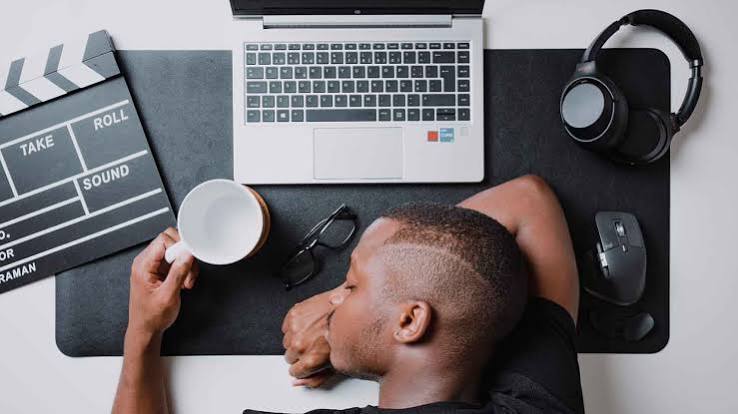 Homem com a cabeça apoiada sobre a mesa de trabalho, ao lado de notebook, fones de ouvido, celular e uma xícara.