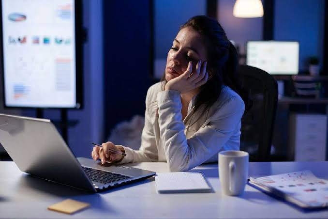 Mulher trabalhando à noite diante de um notebook e monitores, com a cabeça apoiada na mão e expressão de fadiga.